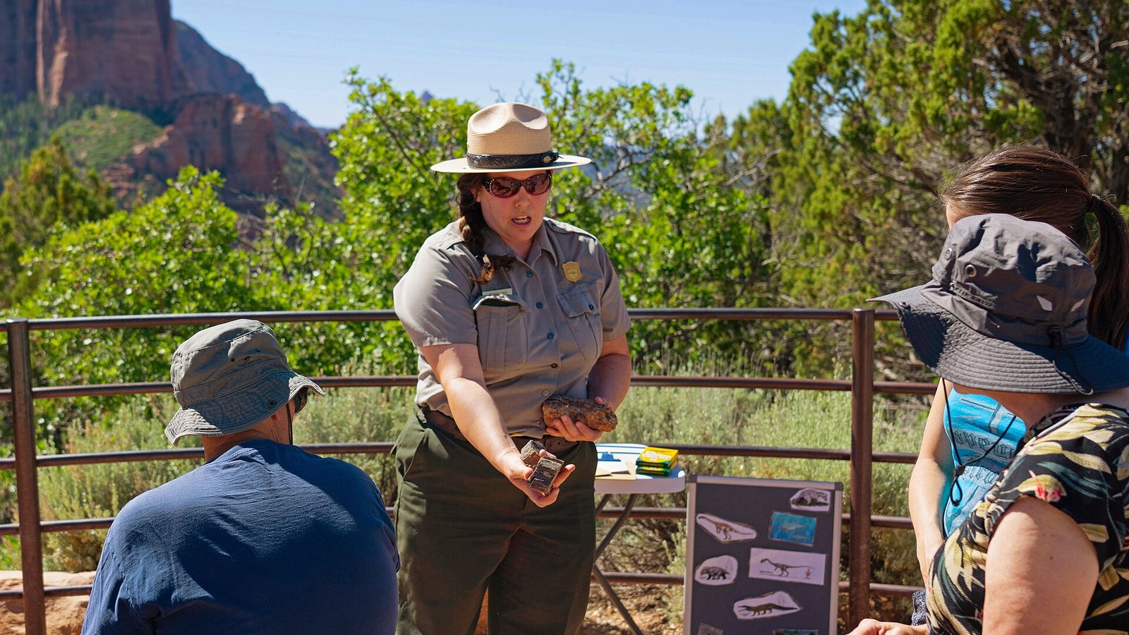 A National Park Service ranger wearing dark sunglasses holds out two fossils for a small crowd of visitors to look at. Ranger Sara gives a t